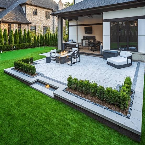 Modern backyard patio with stone pavement, outdoor furniture, fire pit, manicured lawn, and contemporary house with large windows
