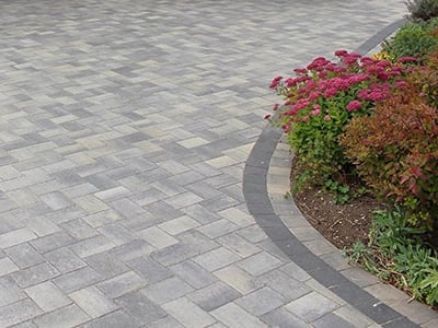 Paved patio with gray brick pattern and curved border edging a landscaped flower bed with red flowering plants