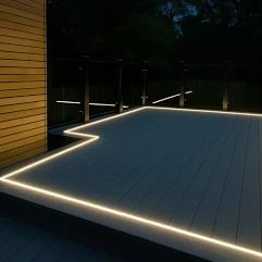 Modern deck with LED strip lighting along edges at night, featuring dark composite decking and glass railings