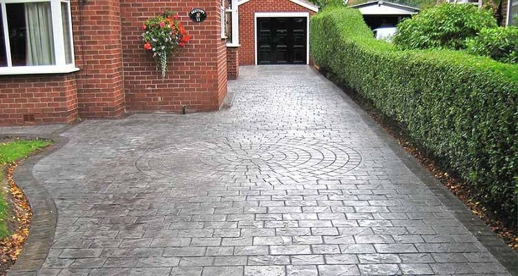 Patterned gray block paved driveway leading to a brick house with garage, bordered by green hedges