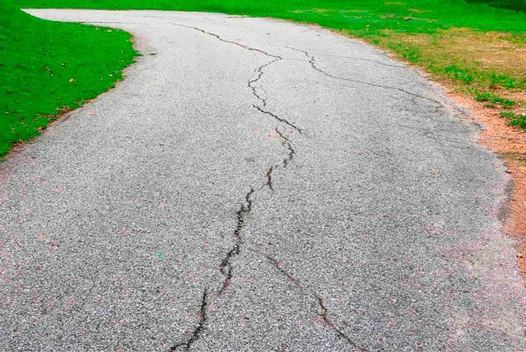 Cracked asphalt pavement with zigzag fissures, bordered by green grass and reddish soil on the sides