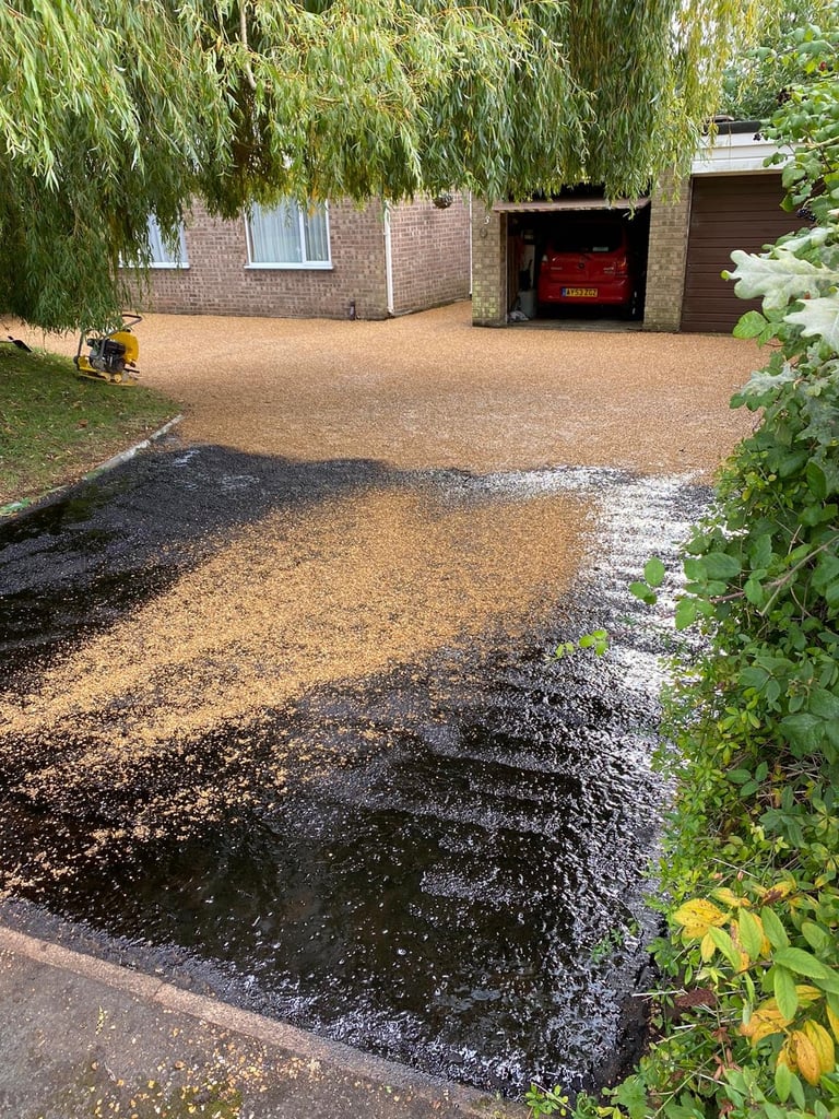 A brick house with gravel driveway, red garage door, weeping willow tree, and wet asphalt section in foreground with green foliage on sides
