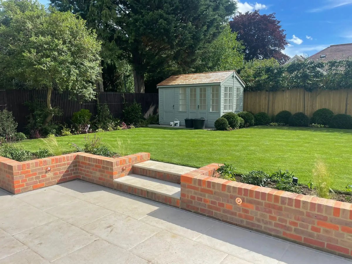Backyard garden with raised red brick beds, patio, green lawn, garden shed, and mature trees under blue sky