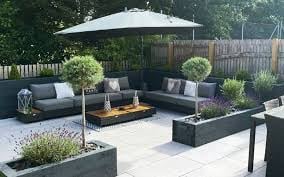 Modern outdoor patio with gray seating, fire pit table, white umbrella, and potted plants on a stone terrace
