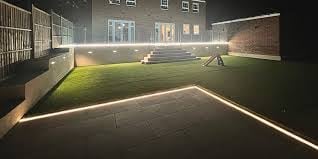 Illuminated outdoor sports court at night with grass field, white boundary lines, and brick building in background