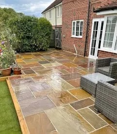 Newly laid patio with mixed stone tiles against a brick house, bordered by green lawn and garden plants