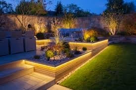 Illuminated modern garden at dusk with warm landscape lighting, raised planter beds, manicured lawn, and trees