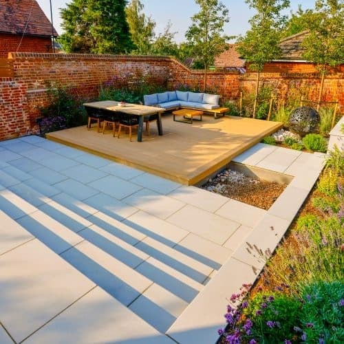 Modern backyard patio with striped pavers, wooden deck, blue sectional sofa, dining table, and brick walls bordered by greenery